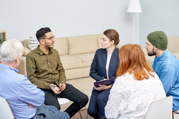 手に話しかける専門性の高い心理学者グループ療法のセッションに参加しながら背景に居心地の良いオフィスのインテリア紙ティッシュで鬱病の混合レース患者 - 写真・画像
