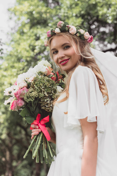 low angle view of beautiful young bride holding wedding bouquet and smiling at camera outdoors - Fotografie, Obrázek