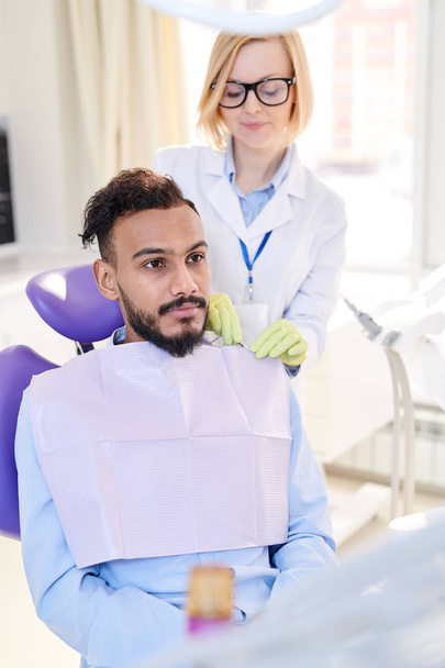Портрет женщины-стоматолога, готовящей молодого человека, сидящего в стоматологическом кресле для лечения зубов в современной стоматологической клинике
 - Фото, изображение