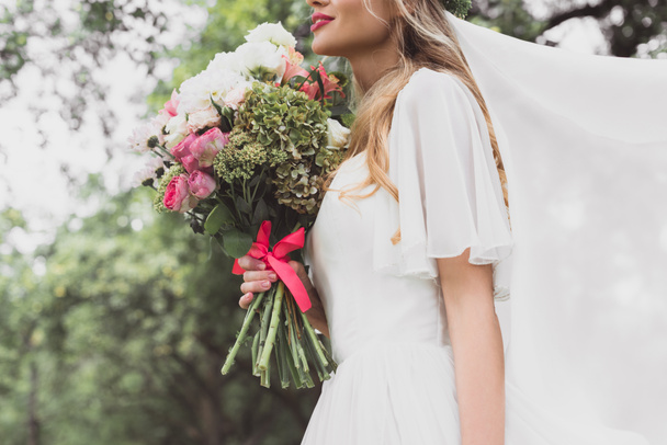 cropped shot of young bride in wedding dress and veil holding beautiful bouquet of flowers - Fotografie, Obrázek