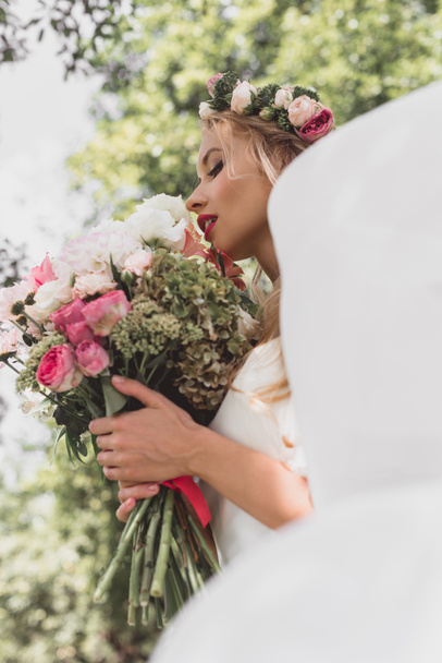 low angle view of tender young bride in floral wreath and veil holding wedding bouquet outdoors - Фото, зображення