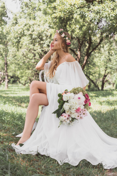 beautiful thoughtful young bride holding wedding bouquet and sitting on chair in park - 写真・画像