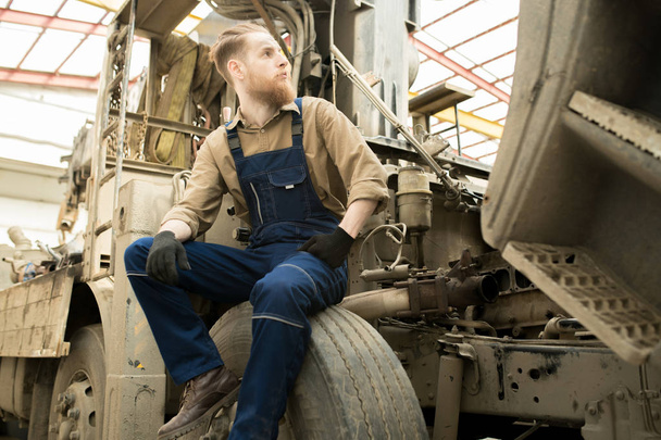 Young bearded male auto technician in overall sitting on wheel of big truck in repair shop and looking to the side - Zdjęcie, obraz