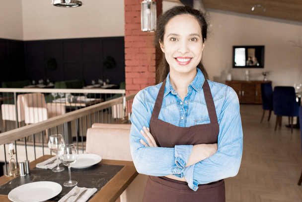 Pozytywny optymistyczny atrakcyjne młody mieszane rasy kelnerka w fartuch przekraczania ręce na klatce piersiowej i uśmiecha się do kamery stojąc w przestronnej restauracji pokój - Zdjęcie, obraz