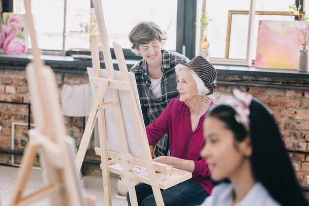 Retrato de una mujer mayor moderna pintando sentada en el caballete en un estudio de arte estudiando arte con una profesora sonriente dando comentarios, escena en un espacioso loft soleado
. - Foto, imagen