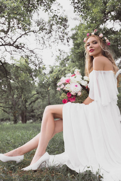 low angle view of beautiful young bride sitting on chair and looking at camera in park - Фото, зображення