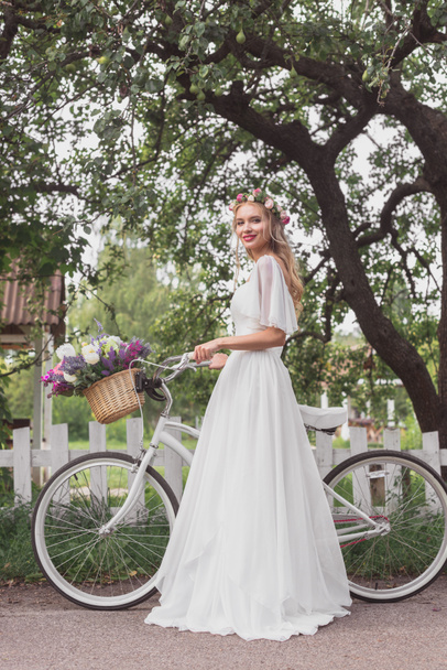 beautiful bride in floral wreath and wedding dress standing with bicycle and smiling at camera  - Fotoğraf, Görsel