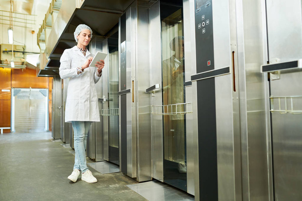 Pracownik fabryki cukiernicze w biały płaszcz, stojący w pobliżu windy i przy użyciu komputera typu tablet.  - Zdjęcie, obraz