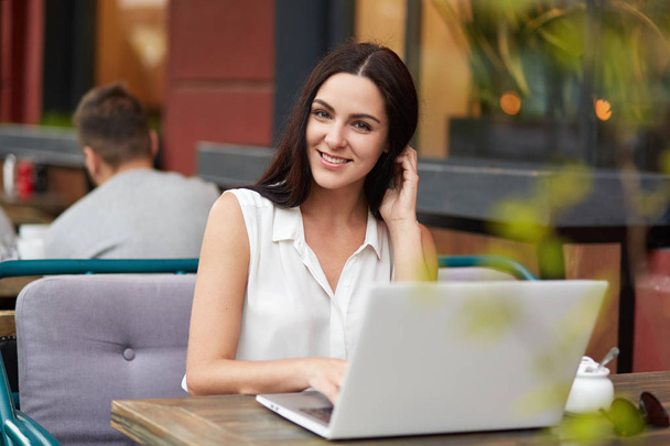 Outdoor shot of pleasant looking brunette young woman with charming smile, spends free time at cozy restaurant, works on laptop computer, searches internet and messages with friends, uses wi fi - Photo, Image
