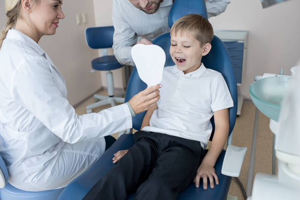 Portrait of happy little boy sitting in dental chair  and looking in mirror showing white teeth during visit to dentist with his father - Photo, Image