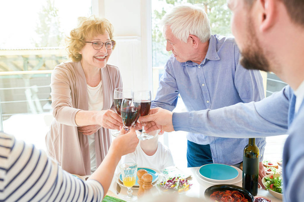 Muotokuva onnellisesta kahden sukupolven perheestä, joka nauttii illallista yhdessä laseja ja paahtoleipää juhlapöydässä ja nauraa loma-juhlan aikana modernissa auringonvalaistussa huoneistossa, kopioi tilaa
 - Valokuva, kuva