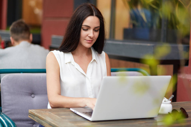 Young beautiful brunette businesswoman dressed in white elegant blouse, sits on outdoor cozy cafeteria, sits in front of opened modern laptop computer, works at business project before meeting - Photo, Image