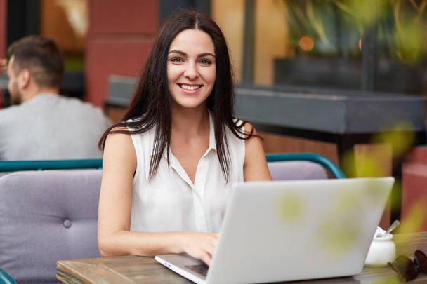 Successful female freelancer works remotely on laptop computer, sits in outdoor cafe, has pleasant smile, enjoys remote work, looks happily at camera. People, leisure and technology concept. - Photo, Image