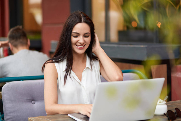 Веселая молодая женщина фрилансер работает на ноутбуке в бистро, имеет зубастую улыбку, как завершает работу, одетый в белую блузку, просматривает интернет-страницу, подключенную к беспроводному Wi-Fi. Технологическая концепция
 - Фото, изображение
