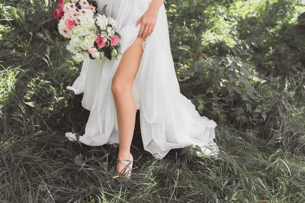 low section of young bride in wedding dress holding bouquet of flowers in garden - Photo, Image