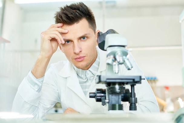 Portret shot van knappe jonge onderzoeker met stijlvolle kapsel aan laboratorium bankje zitten en kijken naar camera, moderne Microscoop op voorgrond - Foto, afbeelding