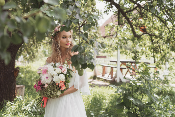 hermosa novia pensativa celebración de ramo de boda y mirando hacia otro lado en el jardín
 - Foto, Imagen