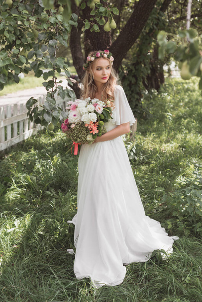 beautiful blonde bride in wedding dress and floral wreath holding bouquet of flowers and looking away - Fotoğraf, Görsel