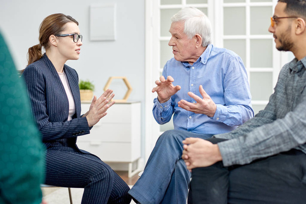 Aged patient wearing denim shirt having productive conversation with highly professional psychologist while participating in group therapy session at cozy office - Photo, Image