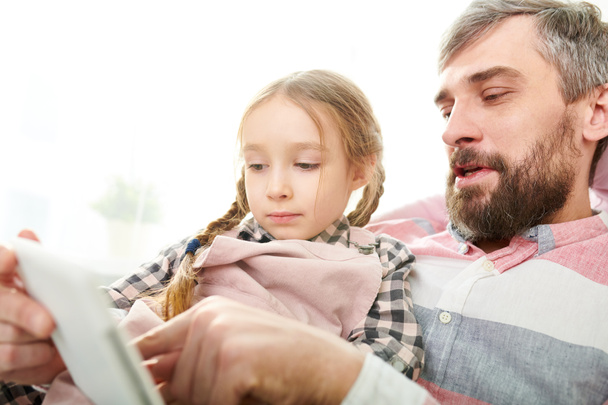 Handsome bearded man and his little daughter playing computer game on digital tablet while spending weekend at home, close-up shot - 写真・画像
