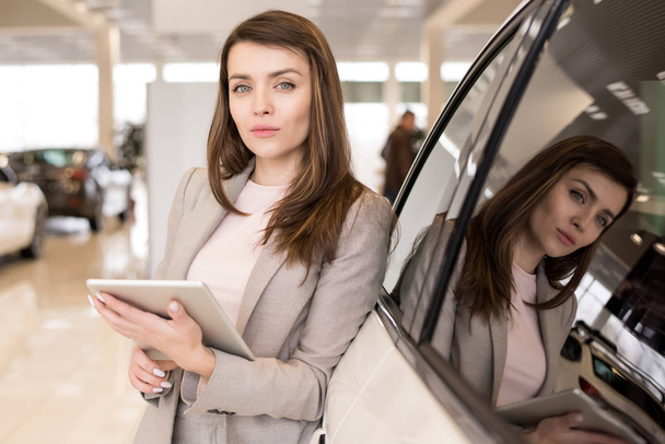 Waist up portrait of beautiful young woman holding clipboard looking at camera standing in luxury dealership showroom - Photo, Image