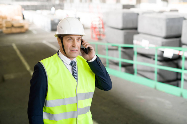 Waist up portrait of mature engineer wearing hardhat and business suit speaking by phone in workshop of modern industrial plant, copy space - Fotoğraf, Görsel