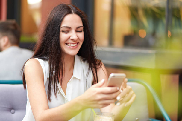 Positive glad female with cheerful expression, holds smart phone, glad to recieve message on smart phone, sits against outdoor cafe interior. Pretty young lady searches social netwoks pages. - Photo, Image