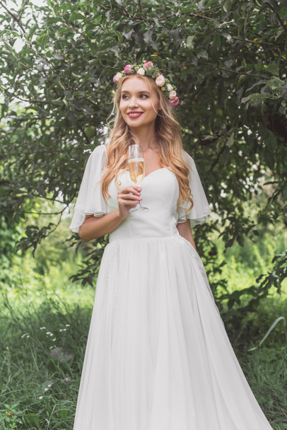beautiful smiling young bride in floral wreath and wedding dress holding glass of champagne and looking away outdoors - Photo, image