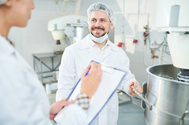 Lächelnder Süsswarenfabrikangestellter steht in weißem Kittel neben Maschinen und schaut Kollege an, der Notizen macht. - Foto, Bild