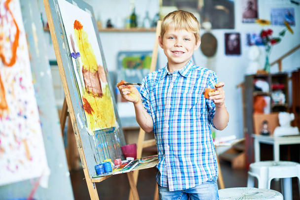 Портрет улыбающегося мальчика, рисующего на мольберте в художественной студии и позирующего, смотрящего в камеру, держащего кисть и краску
 - Фото, изображение