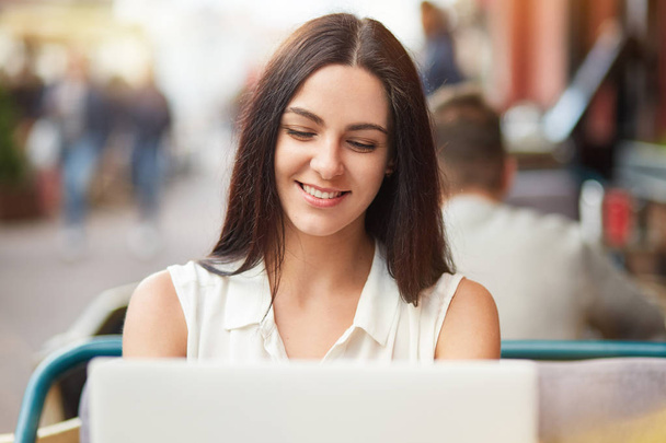 horizontale Aufnahme der schönen jungen kaukasischen Frau sitzt vor geöffnetem Laptop, lächelt sanft, Nachrichten in sozialen Netzwerken, verbunden mit drahtlosem Interent. Menschen und Lebensstil. - Foto, Bild
