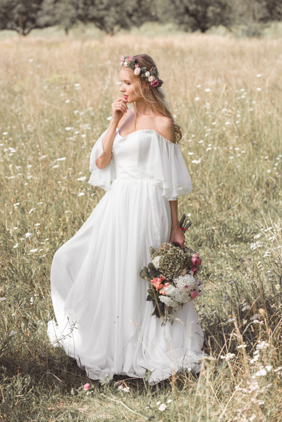 beautiful pensive bride in wedding dress and floral wreath holding bouquet of flowers in field - Foto, afbeelding