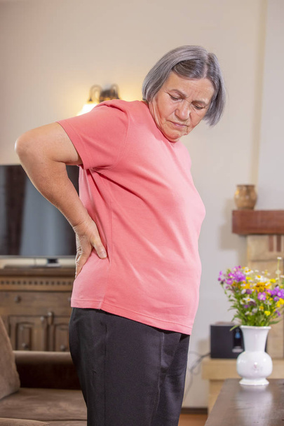 Mature woman suffering from backache at home. Massaging lower back with hand, feeling exhausted, standing in living room. - Foto, Imagen