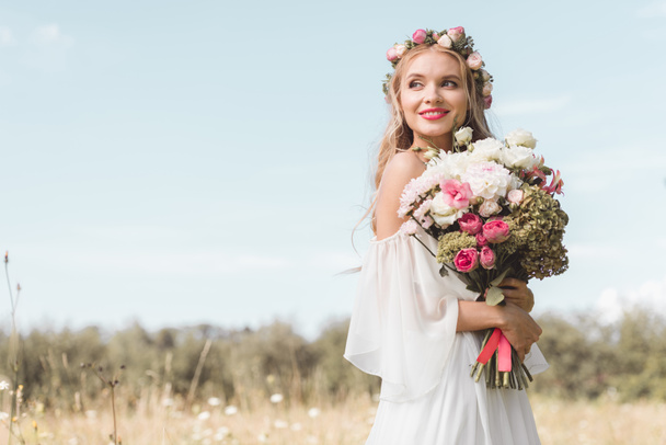 beautiful happy young bride holding bouquet of flowers and looking away outdoors  - Фото, изображение