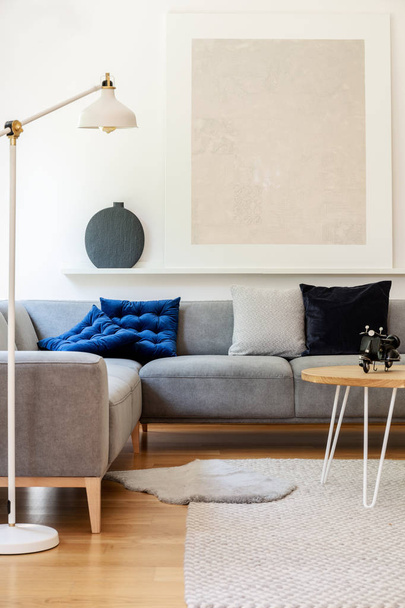 Real photo of bright living room interior with simple poster, metal lamp, grey corner couch and carpet - Valokuva, kuva