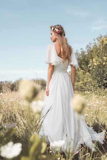 back view of beautiful young bride in wedding dress and floral wreath walking on field - Foto, imagen