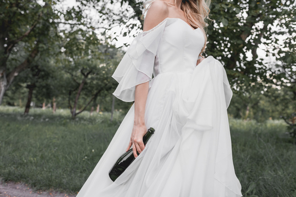 Schnappschuss einer jungen Braut im Brautkleid, die eine Flasche Wein hält und im Park spaziert - Foto, Bild