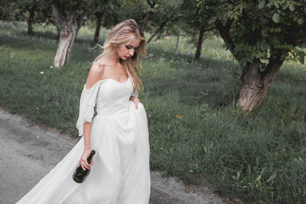 засмучена молода наречена у весільній сукні тримає пляшку вина і гуляє в парку
  - Фото, зображення