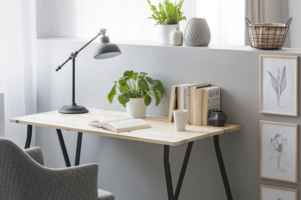 Real photo of wooden desk with fresh plant, black lamp, coffee cup and books standing on half-wall with simple posters - Φωτογραφία, εικόνα