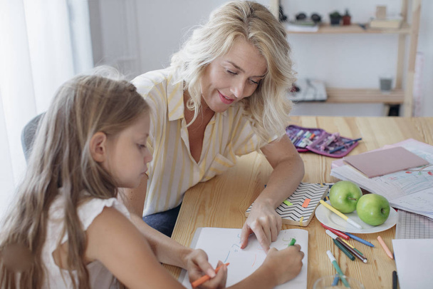 Beautiful blonde caucasian girl studying at home with her mom helping. - Photo, image