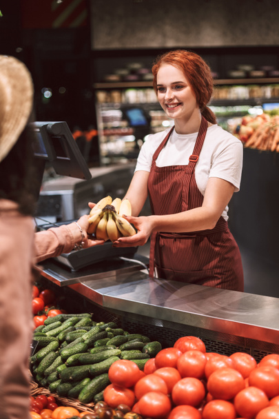 近代的なスーパー マーケットの顧客にバナナを与えて喜んで野菜とカウンターの後ろにエプロンの若い笑顔販売 - 写真・画像
