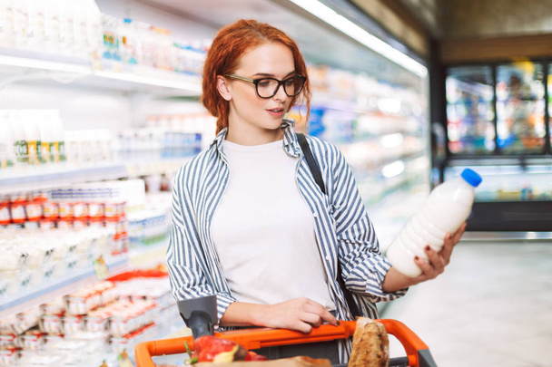 Linda chica en gafas graduadas y camisa a rayas con carrito de compras cuidadosamente mirando en botella de leche en las manos en el supermercado moderno
 - Foto, Imagen