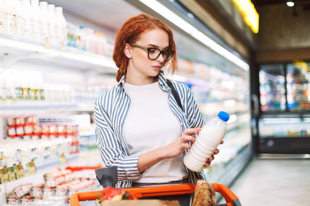 Mujer joven en gafas graduadas y camisa a rayas con carrito de compras mirando cuidadosamente en botella de leche en el supermercado moderno
 - Foto, Imagen