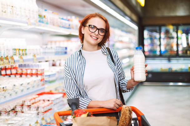 Chica alegre en gafas y camisa a rayas con carrito de compras mirando felizmente en cámara sosteniendo botella de leche en la mano en el supermercado moderno
 - Foto, Imagen