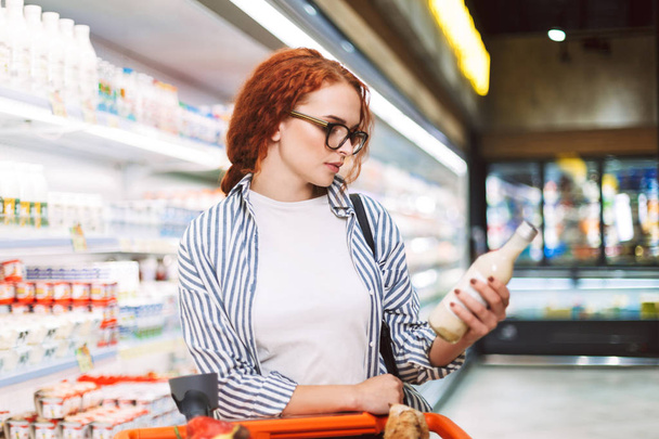 Giovane donna pensierosa in occhiali e camicia a righe con carrello della spesa pensieroso guardando sulla bottiglia di latte nel supermercato moderno
 - Foto, immagini