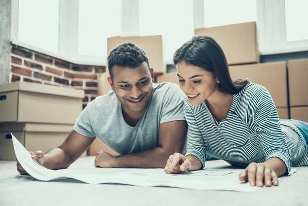 Young Smiling Couple Enjoy Moving in New House. Young Smiling Couple with Cardboard Boxes on background lying on Floor and look at Project of New House. Relocation and House Moving Concept - Foto, Imagem