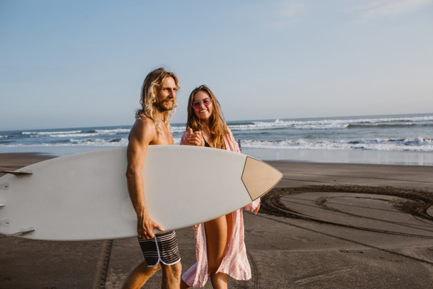 happy couple walking together on beach with surfing board in bali, indonesia - Φωτογραφία, εικόνα
