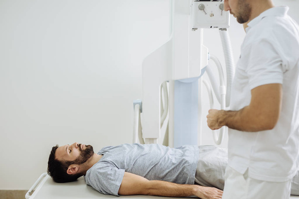 Fiatal orvosi technikus működő egy röntgengép, miközben a beteg fekszik előtte a klinikán a ágy. - Fotó, kép