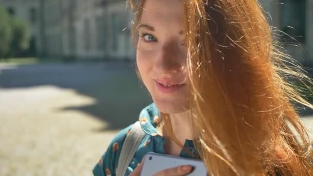 Beautiful young ginger woman holding tablet and looking at camera, standing on street during sunny day, happy female smiling - Séquence, vidéo