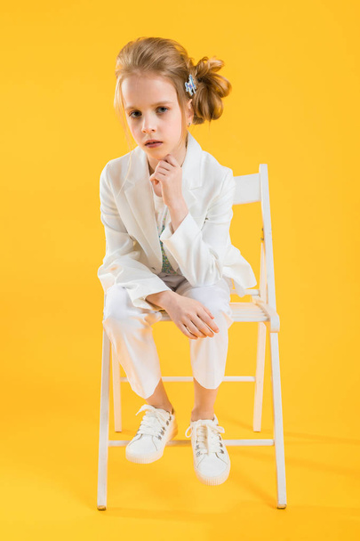 Dospívající dívka v bílých šatech sedí na židli na žlutém podkladu. - Fotografie, Obrázek